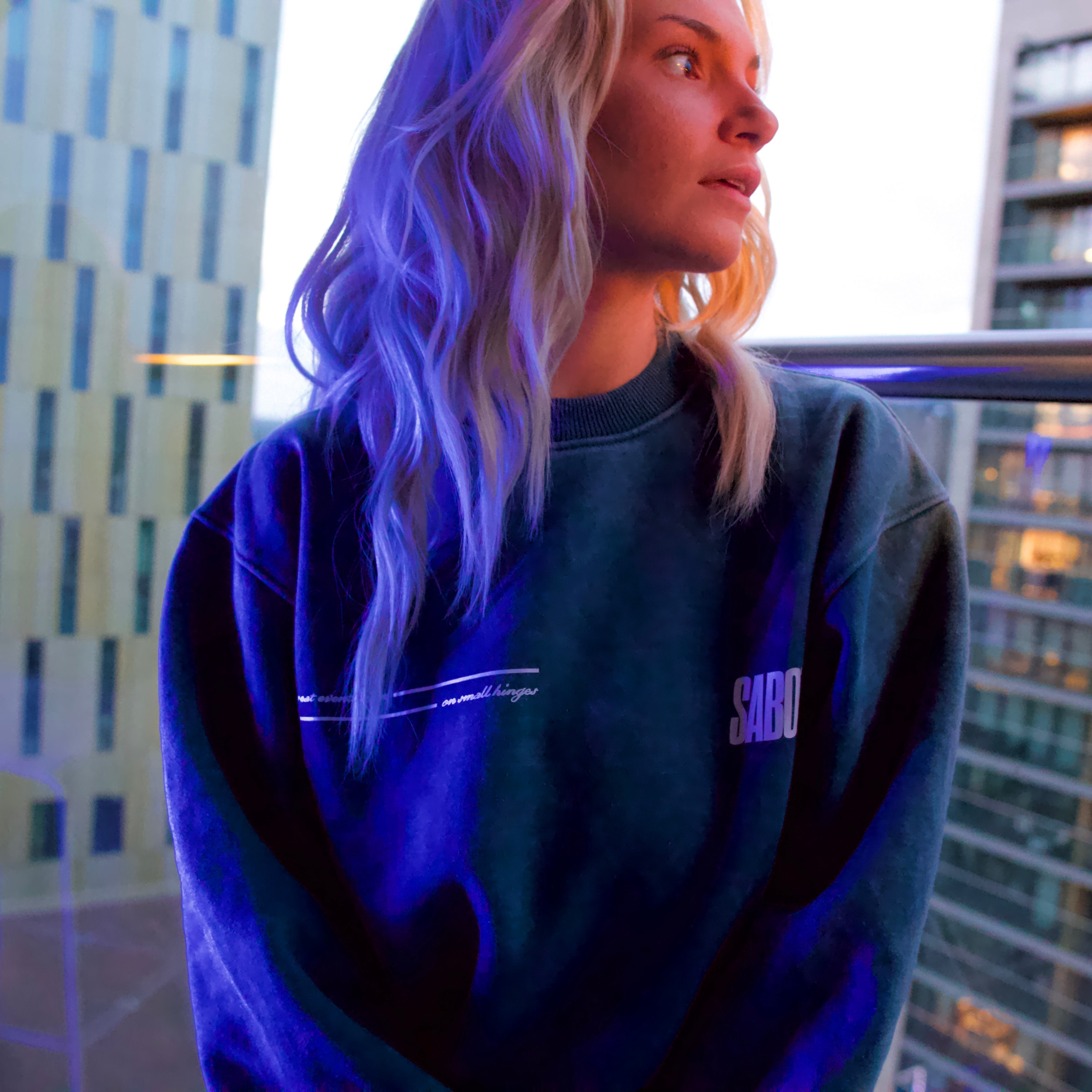 Person wearing a dark blue sweatshirt with 'SABOTAGE' logo, standing on a balcony with cityscape background