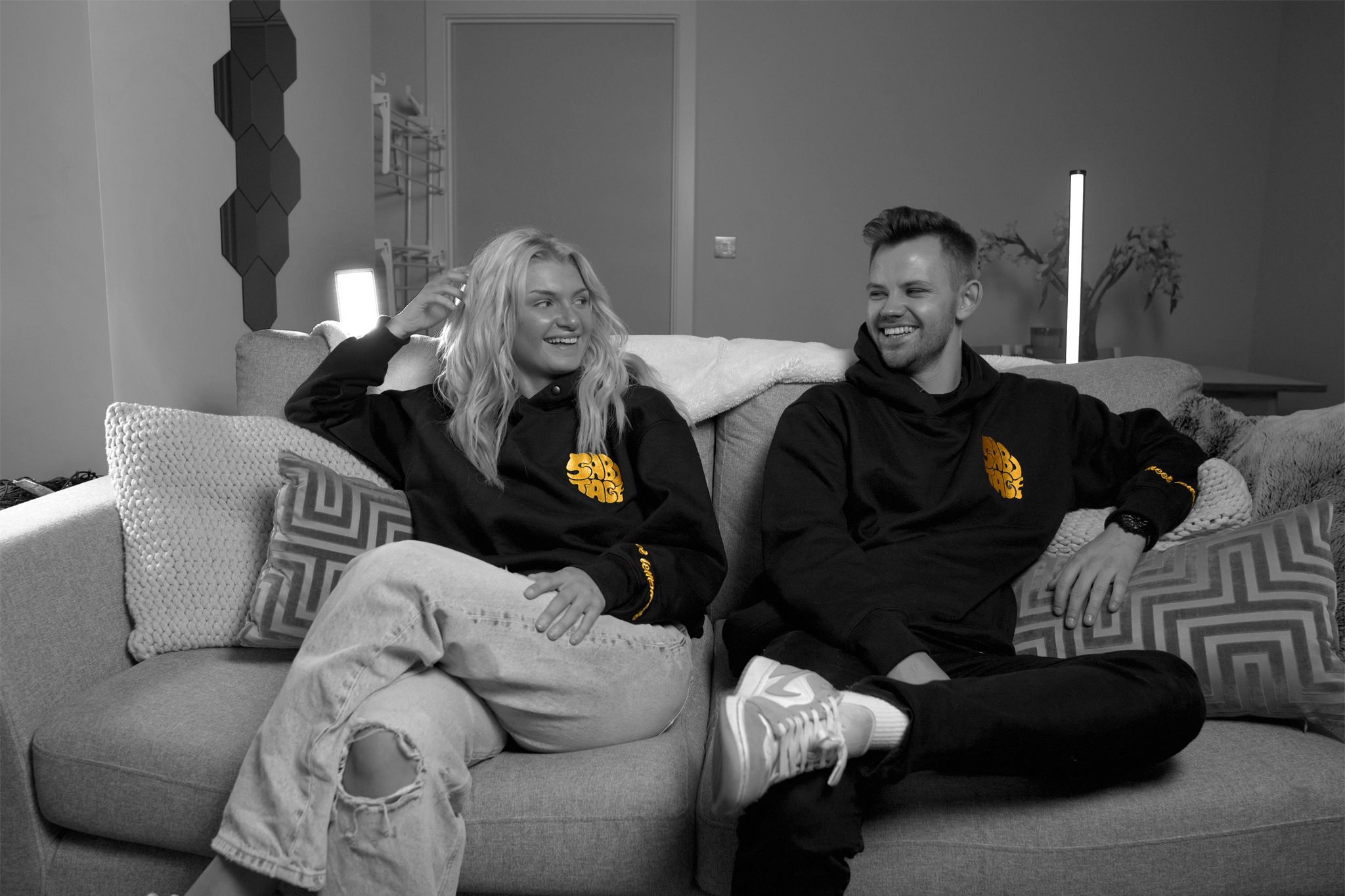 Two people, a man and a woman sitting on a sofa wearing Black hoodies with a bold yellow embroidered design on the cuff and chest