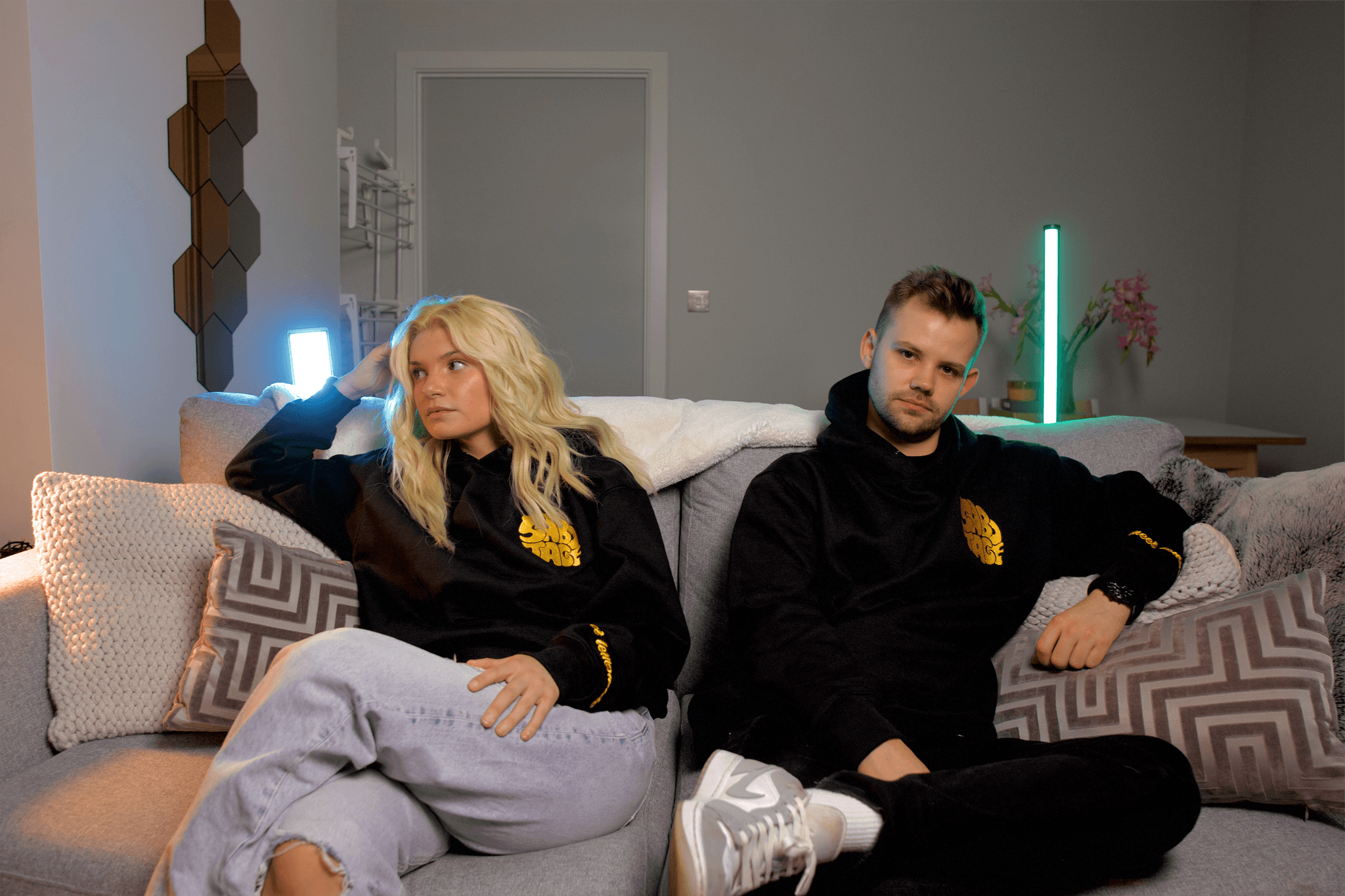 Two people, a man and a woman sitting on a sofa wearing Black hoodies with a bold yellow embroidered design on the cuff and chest
